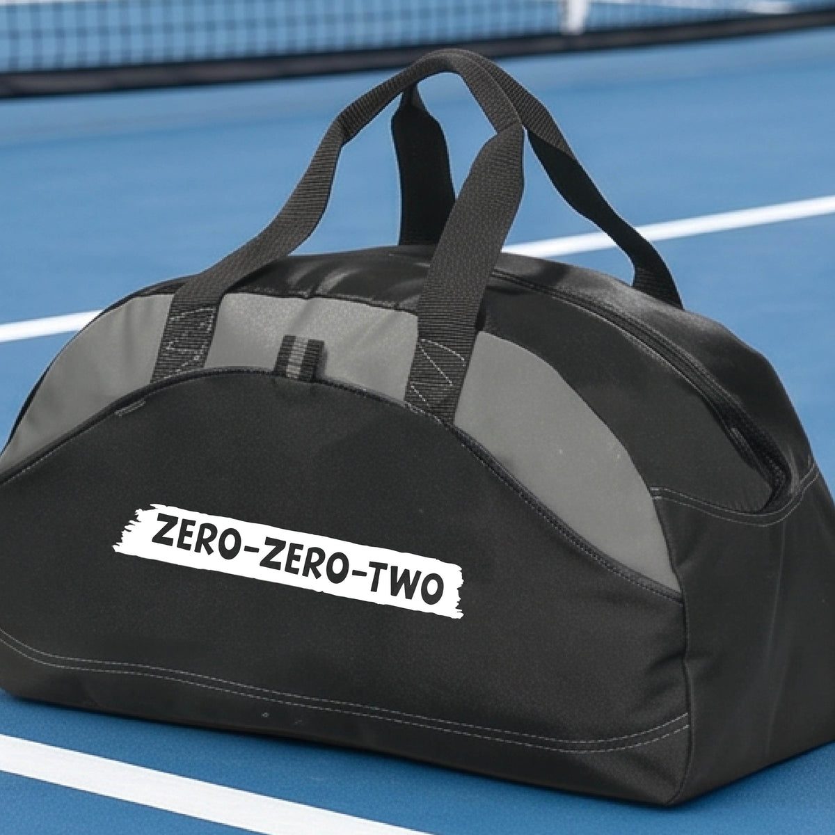 A close-up of a black duffel bag with gray panels and a white graphic of artistic banner with the cutout words of ‘ZERO ZERO TWO’, on the front. 