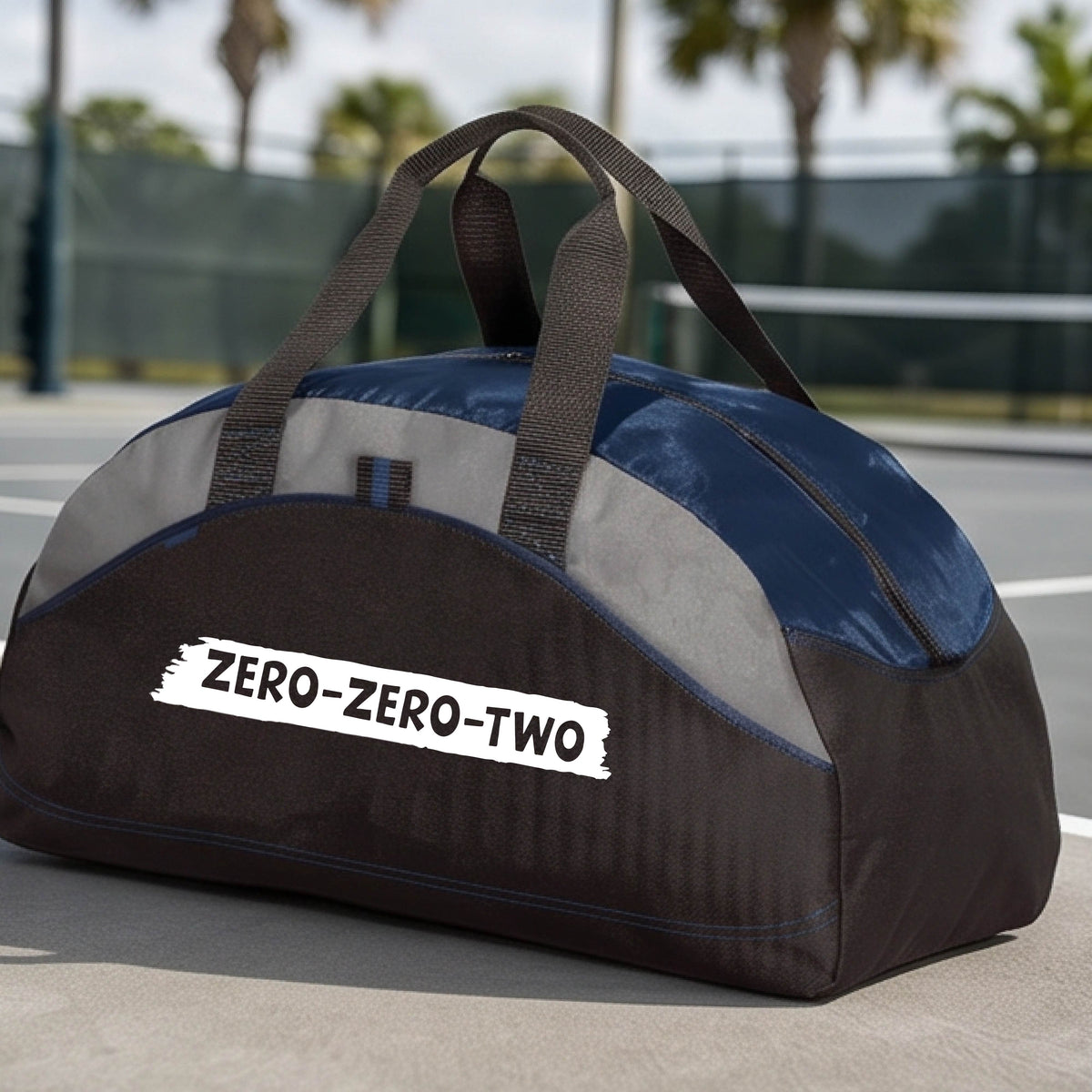A close-up of a navy blue and black duffel bag with gray panels and a white graphic of artistic banner with the cutout words of ‘ZERO ZERO TWO’, on the front. 