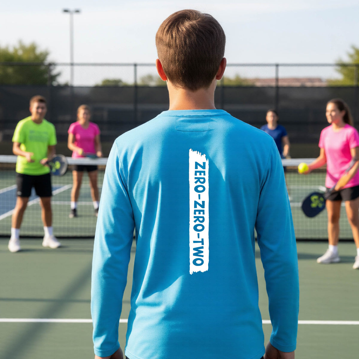 A close-up of a young person on a pickleball wearing a bright blue short-sleeve t-shirt, , with a white graphic of artistic banner with the cutout words of ‘ZERO ZERO TWO’ , on the back of the shirt. 