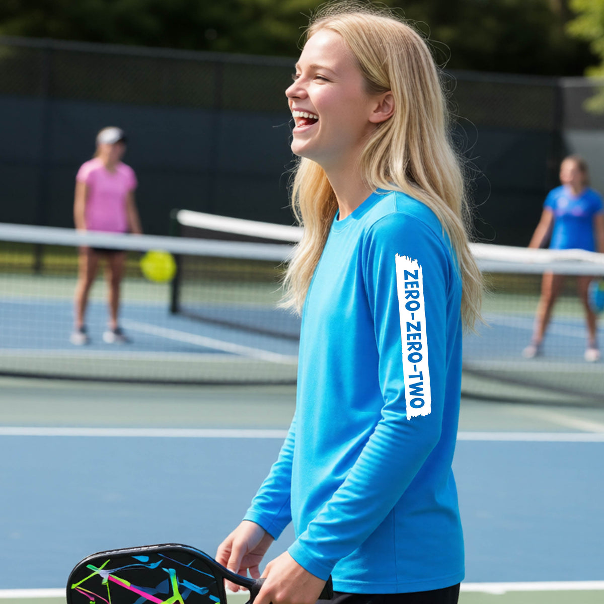 A close-up of a young person on a pickleball wearing a bright blue short-sleeve t-shirt, , with a white graphic of artistic banner with the cutout words of ‘ZERO ZERO TWO’ , on the sleeve of the shirt. 
