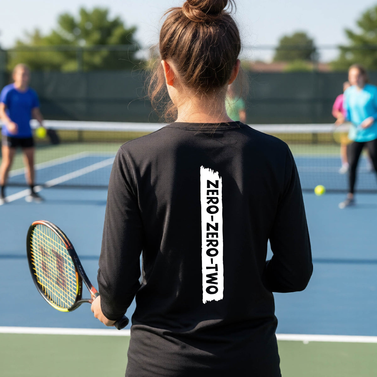 A close-up of a young person on a pickleball wearing a black short-sleeve t-shirt, , with a white graphic of artistic banner with the cutout words of ‘ZERO ZERO TWO’ , on the back of the shirt. 