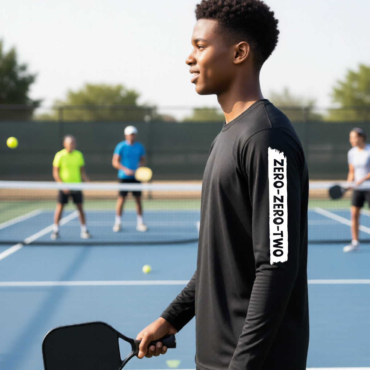 A close-up of a young person on a pickleball wearing a black short-sleeve t-shirt, , with a white graphic of artistic banner with the cutout words of ‘ZERO ZERO TWO’ , on the sleeve of the shirt. 