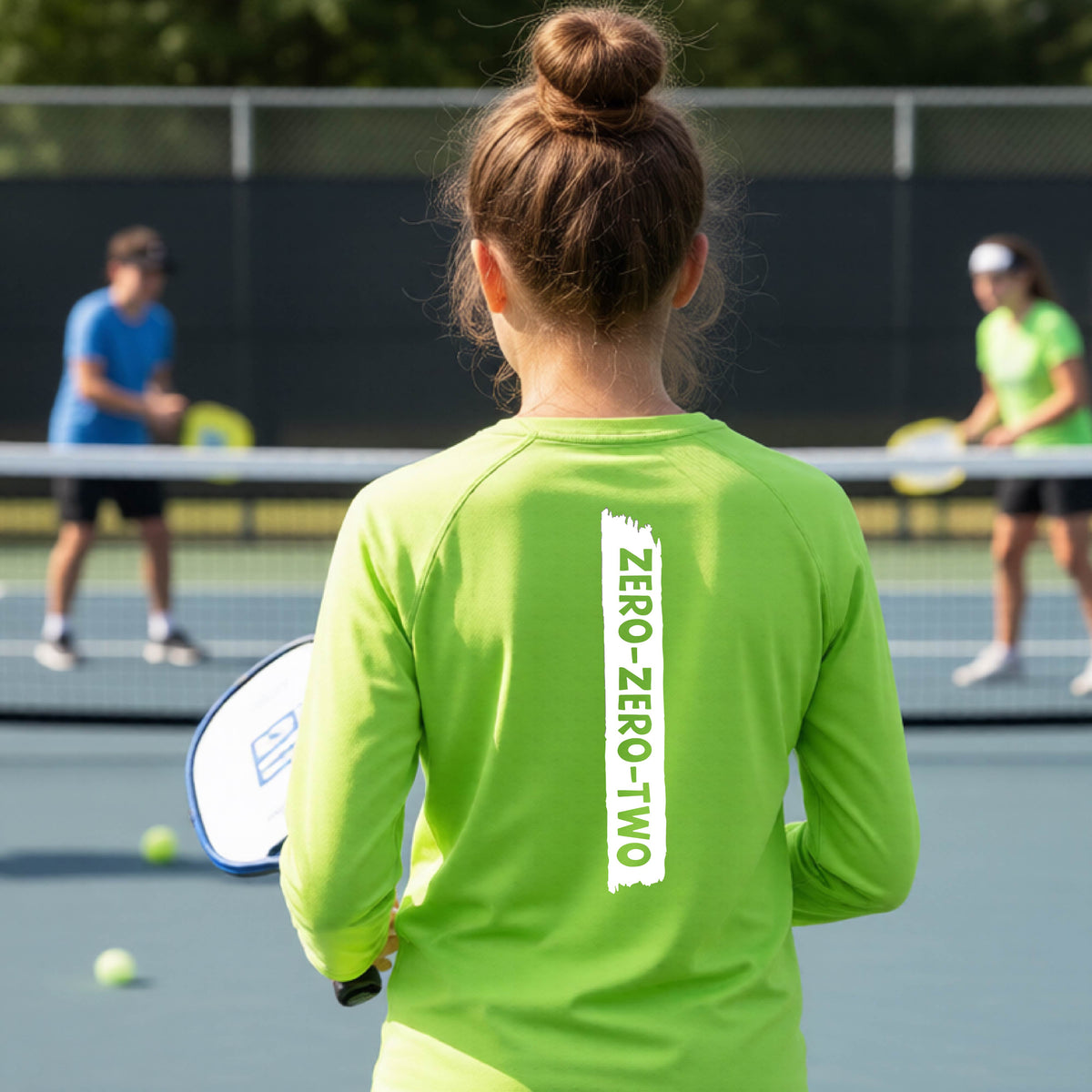 A close-up of a young person on a pickleball wearing a lime green short-sleeve t-shirt, , with a white graphic of artistic banner with the cutout words of ‘ZERO ZERO TWO’ , on the back of the shirt. 