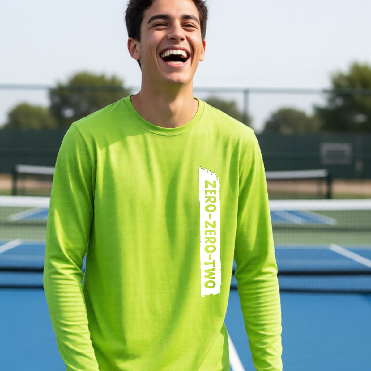 A close-up of a young person on a pickleball wearing a lime green short-sleeve t-shirt, , with a white graphic of artistic banner with the cutout words of ‘ZERO ZERO TWO’ , on the front of the shirt. 