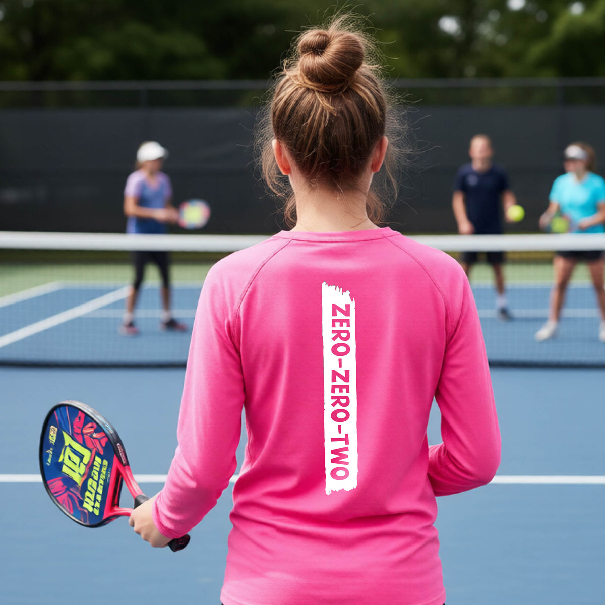 A close-up of a young person on a pickleball wearing a pink short-sleeve t-shirt, , with a white graphic of artistic banner with the cutout words of ‘ZERO ZERO TWO’ , on the back of the shirt.