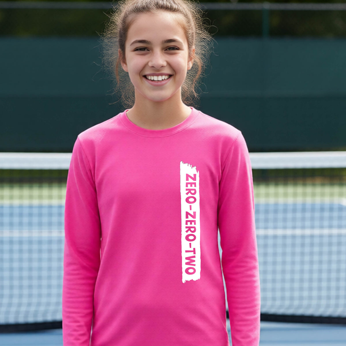 A close-up of a young person on a pickleball wearing a pink short-sleeve t-shirt, , with a white graphic of artistic banner with the cutout words of ‘ZERO ZERO TWO’ , on the front of the shirt. 
