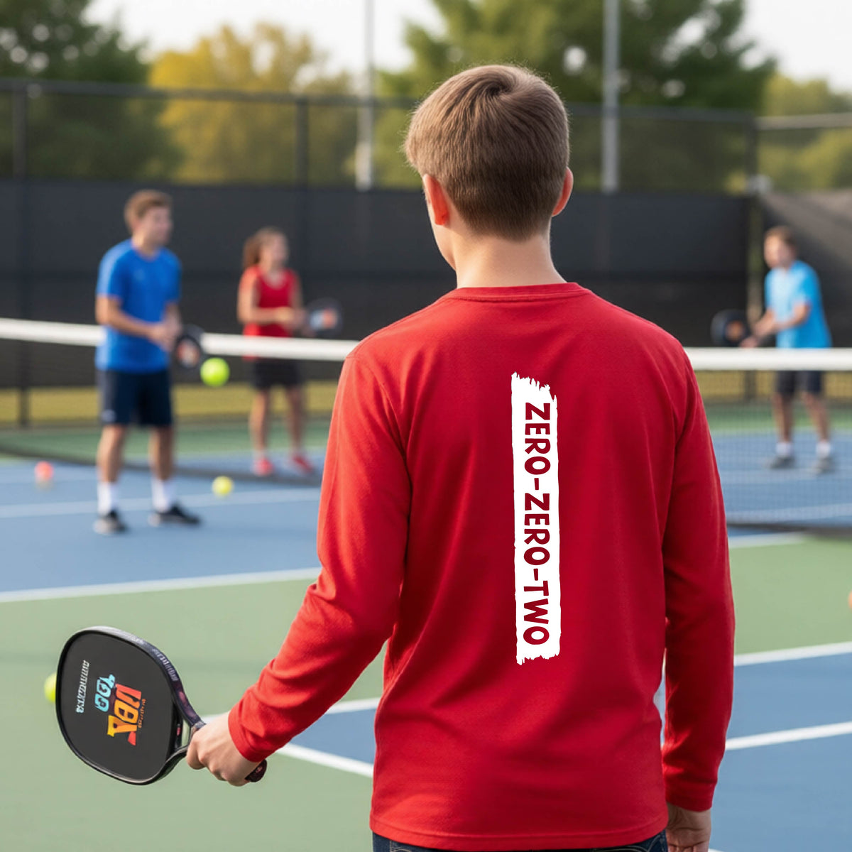 A close-up of a young person on a pickleball wearing a red short-sleeve t-shirt, , with a white graphic of artistic banner with the cutout words of ‘ZERO ZERO TWO’ , on the back of the shirt. 
