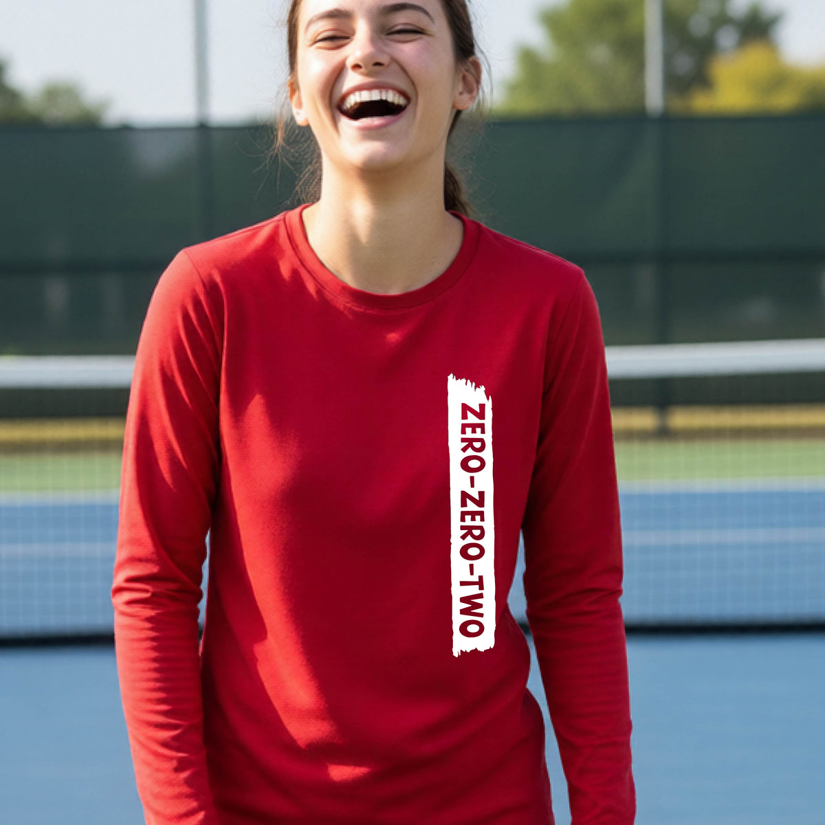 A close-up of a young person on a pickleball wearing a red short-sleeve t-shirt, , with a white graphic of artistic banner with the cutout words of ‘ZERO ZERO TWO’ , on the sleeve shirt. 