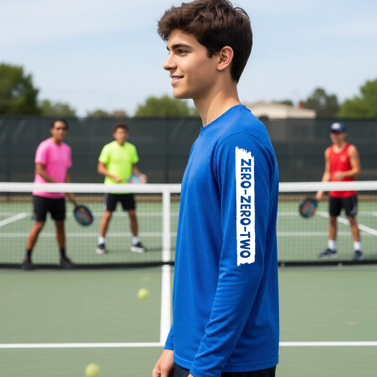 A close-up of a young person on a pickleball wearing a royal blue short-sleeve t-shirt, , with a white graphic of artistic banner with the cutout words of ‘ZERO ZERO TWO’ , on the sleeve of the shirt. 