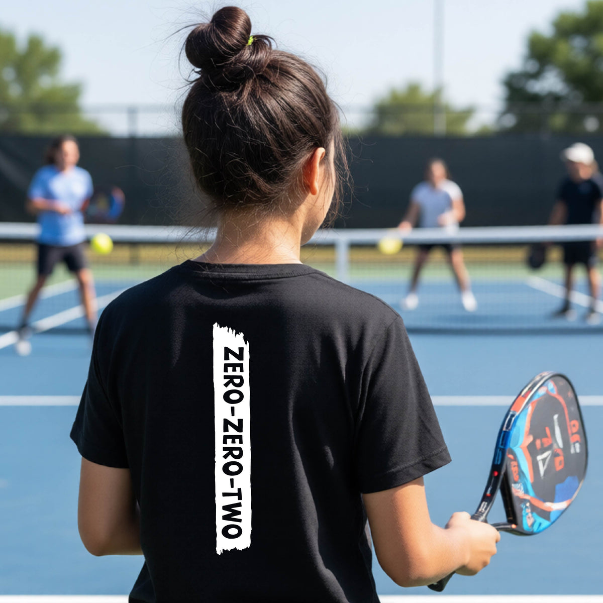 A close-up of a young person on a pickleball wearing a black short-sleeve t-shirt, , with a white graphic of artistic banner with the cutout words of ‘ZERO ZERO TWO’ , on the back of the shirt. 