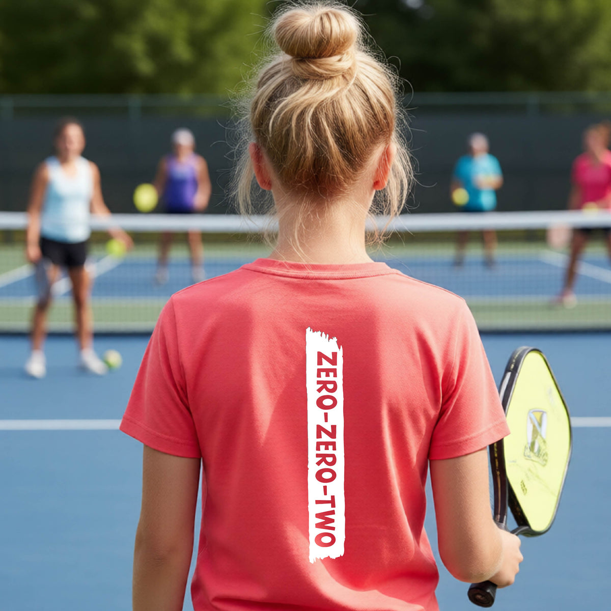 A close-up of a young person on a pickleball wearing a coral pink short-sleeve t-shirt, , with a white graphic of artistic banner with the cutout words of ‘ZERO ZERO TWO’ , on the back of the shirt. 