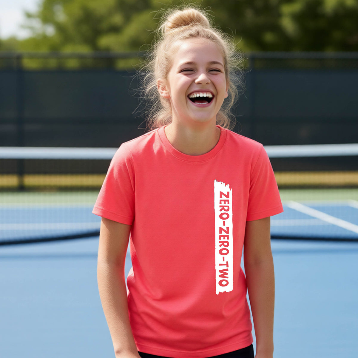 A close-up of a young person on a pickleball wearing a coral pink short-sleeve t-shirt, , with a white graphic of artistic banner with the cutout words of ‘ZERO ZERO TWO’ , on the front of the shirt. 