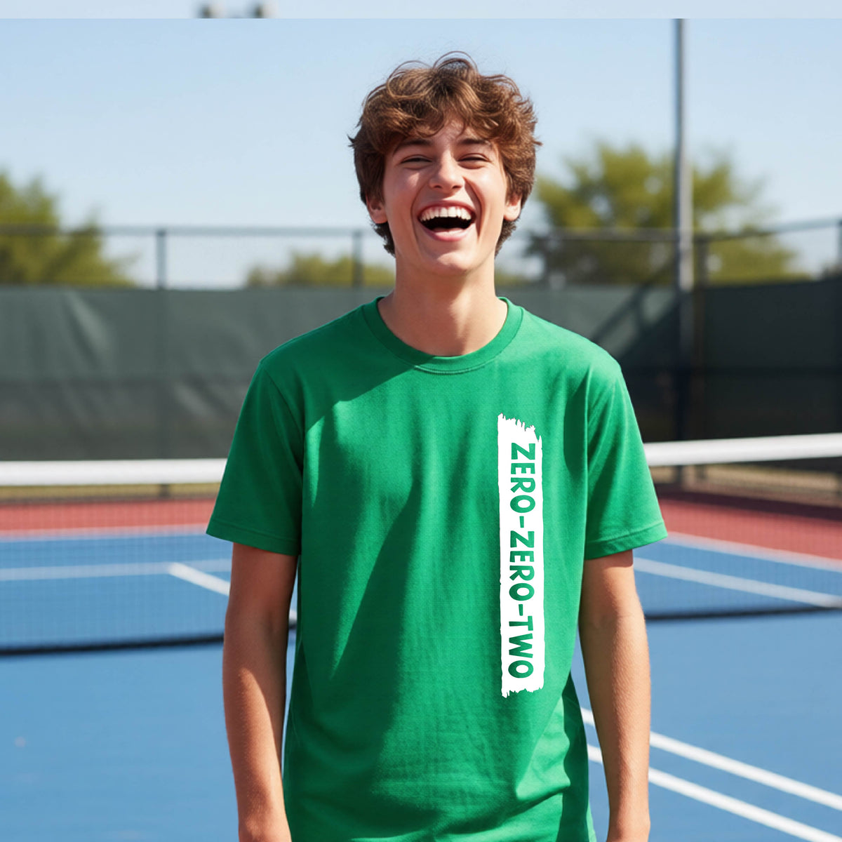 A close-up of a young person on a pickleball wearing a green short-sleeve t-shirt, , with a white graphic of artistic banner with the cutout words of ‘ZERO ZERO TWO’ , on the front of the shirt. 