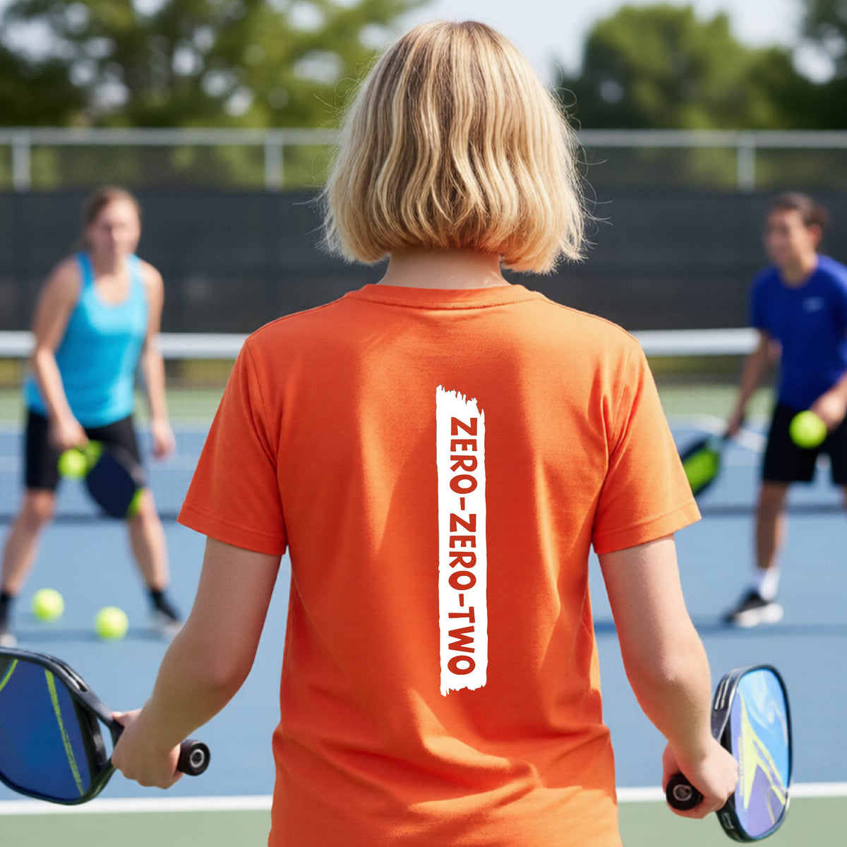 A close-up of a young person on a pickleball wearing an orange short-sleeve t-shirt, , with a white graphic of artistic banner with the cutout words of ‘ZERO ZERO TWO’ , on the shirt. 