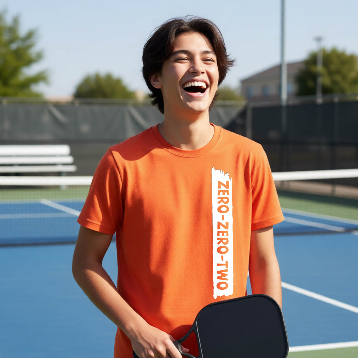 A close-up of a young person on a pickleball wearing an orange short-sleeve t-shirt, , with a white graphic of artistic banner with the cutout words of ‘ZERO ZERO TWO’ , on the front of the shirt. 