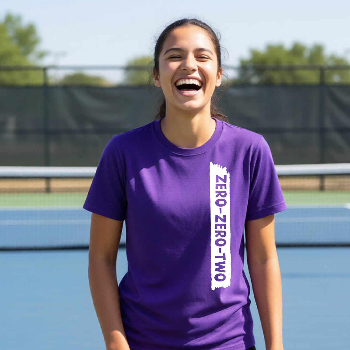 A close-up of a young person on a pickleball wearing an purple short-sleeve t-shirt, , with a white graphic of artistic banner with the cutout words of ‘ZERO ZERO TWO’ , on the front of the shirt. 