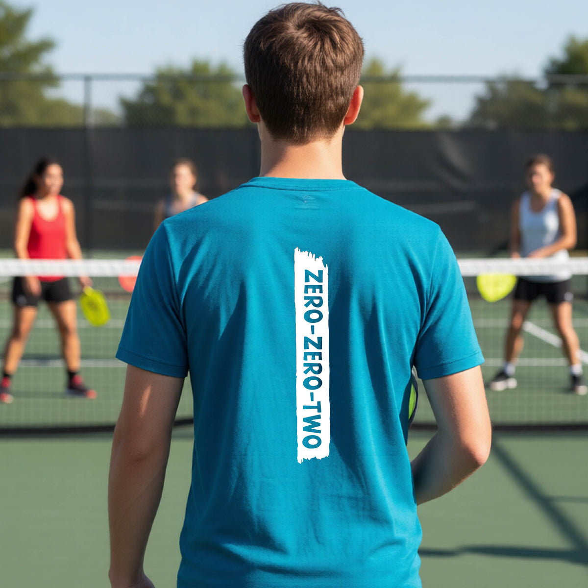 A close-up of a young person on a pickleball wearing an teal short-sleeve t-shirt, , with a white graphic of artistic banner with the cutout words of ‘ZERO ZERO TWO’ , on the back of the shirt. 
