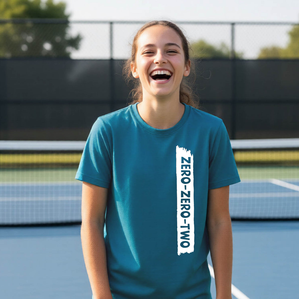 A close-up of a young person on a pickleball wearing an teal short-sleeve t-shirt, , with a white graphic of artistic banner with the cutout words of ‘ZERO ZERO TWO’ , on the front of the shirt. 