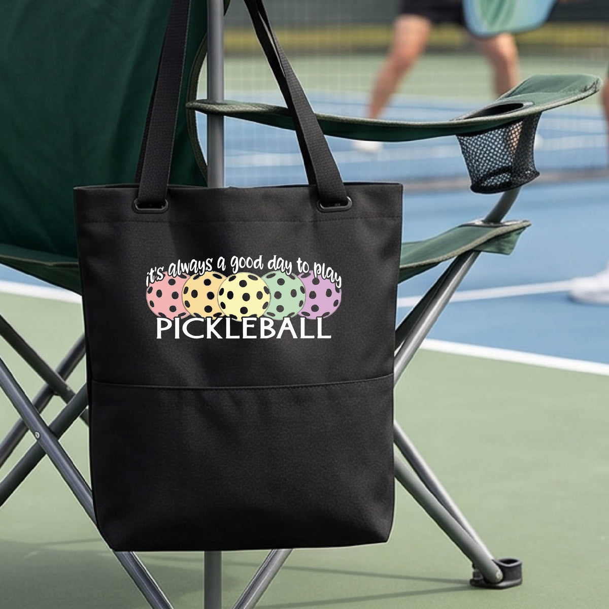 A black tote bag with a colorful pickleball graphic and the text 'it's always a good day to play PICKLEBALL' hanging from a chair on a pickleall court. 