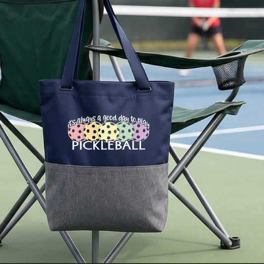 A navy blue and gray tote bag with a colorful pickleball graphic and the text 'it's always a good day to play PICKLEBALL' hanging from a chair on a pickleall court. 