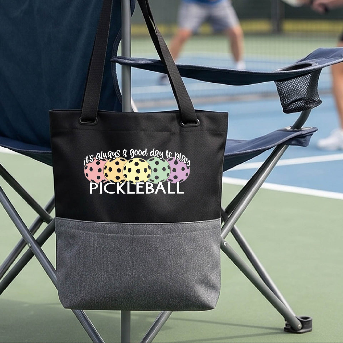 A black and gray tote bag with a colorful pickleball graphic and the text 'it's always a good day to play PICKLEBALL' hanging from a chair on a pickleall court. 