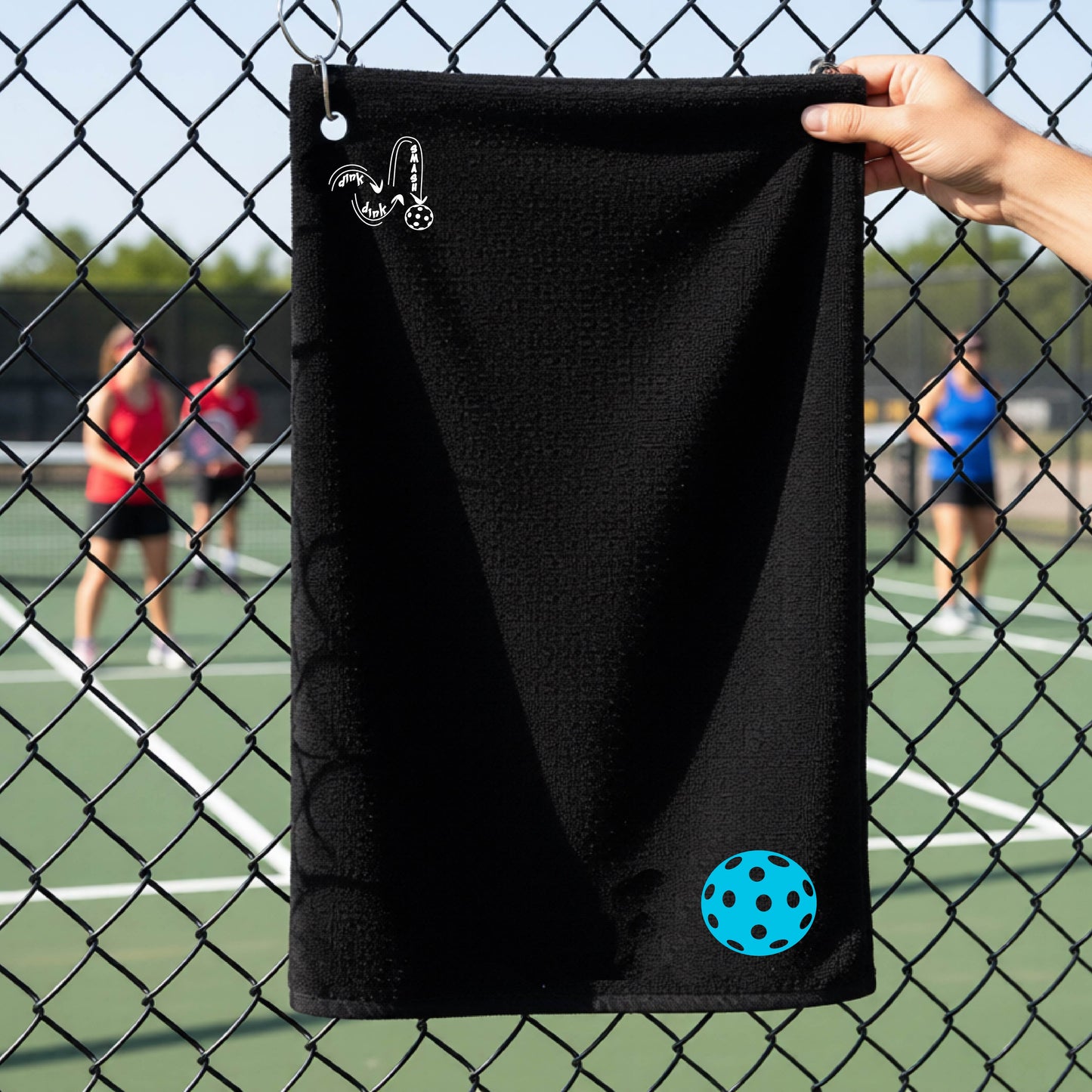 A black terry cloth towel is held up in front of a chain-link fence at a pickleall court. The towel features a silver metal grommet and hook at the top, with the text"DINK DINK SMASH" and a colorful pickleball graphic near the bottom right.