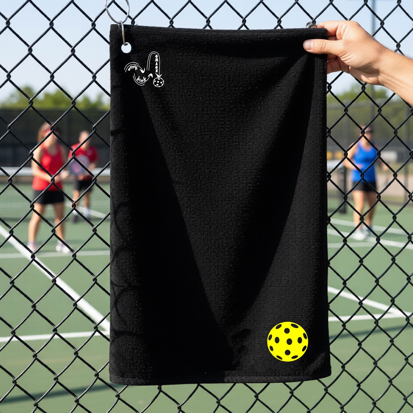 A black terry cloth court towel is held up in front of a chain-link fence at a pickleall court. The towel features a silver metal grommet and hook at the top, with the text"DINK DINK SMASH" and a colorful pickleball graphic near the bottom right.