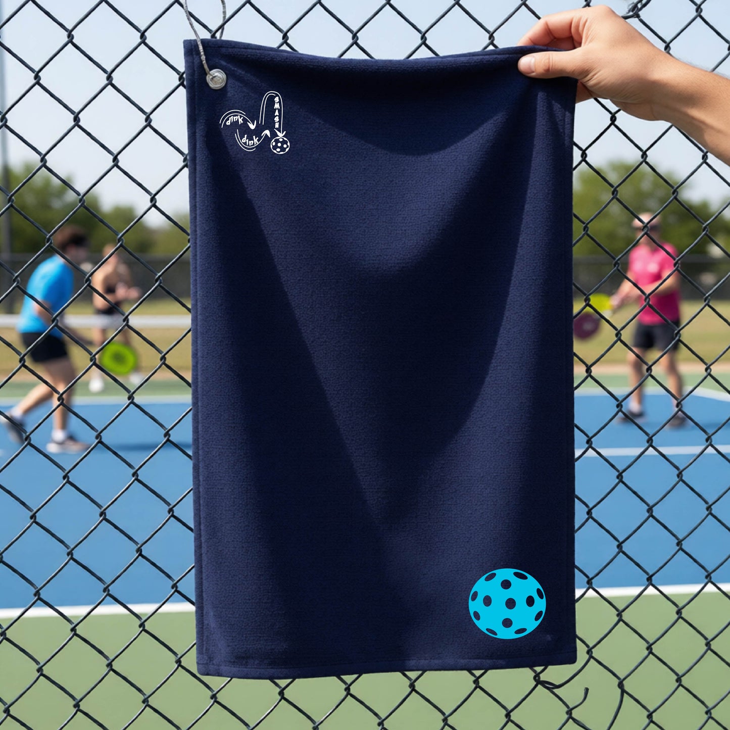 A navy blue terry cloth court towel is held up in front of a chain-link fence at a pickleall court. The towel features a silver metal grommet and hook at the top, with the text"DINK DINK SMASH" and a colorful pickleball graphic near the bottom right.