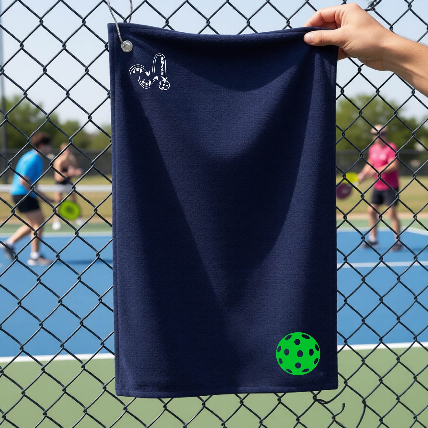 A navy blue terry cloth court towel is held up in front of a chain-link fence at a pickleall court. The towel features a silver metal grommet and hook at the top, with the text"DINK DINK SMASH" and a colorful pickleball graphic near the bottom right.