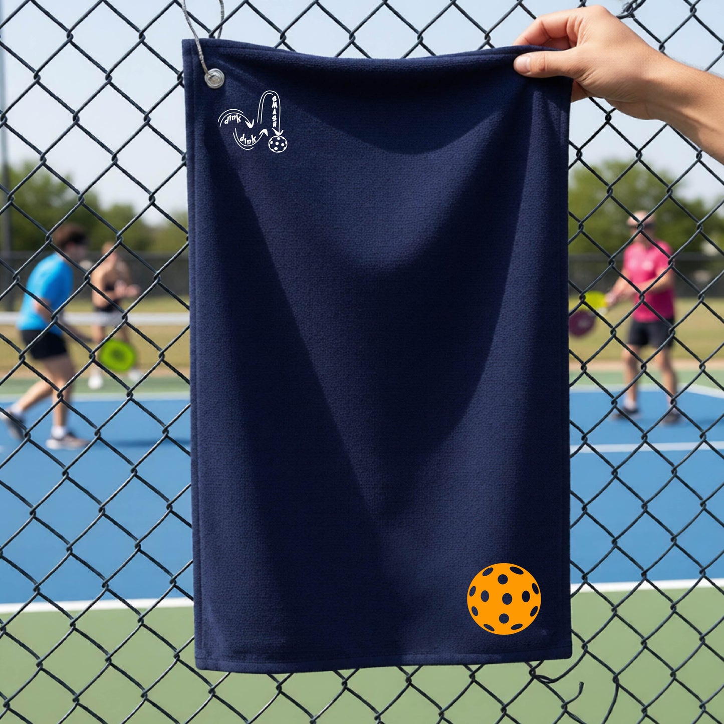 A navy blue terry cloth court towel is held up in front of a chain-link fence at a pickleall court. The towel features a silver metal grommet and hook at the top, with the text"DINK DINK SMASH" and a colorful pickleball graphic near the bottom right.