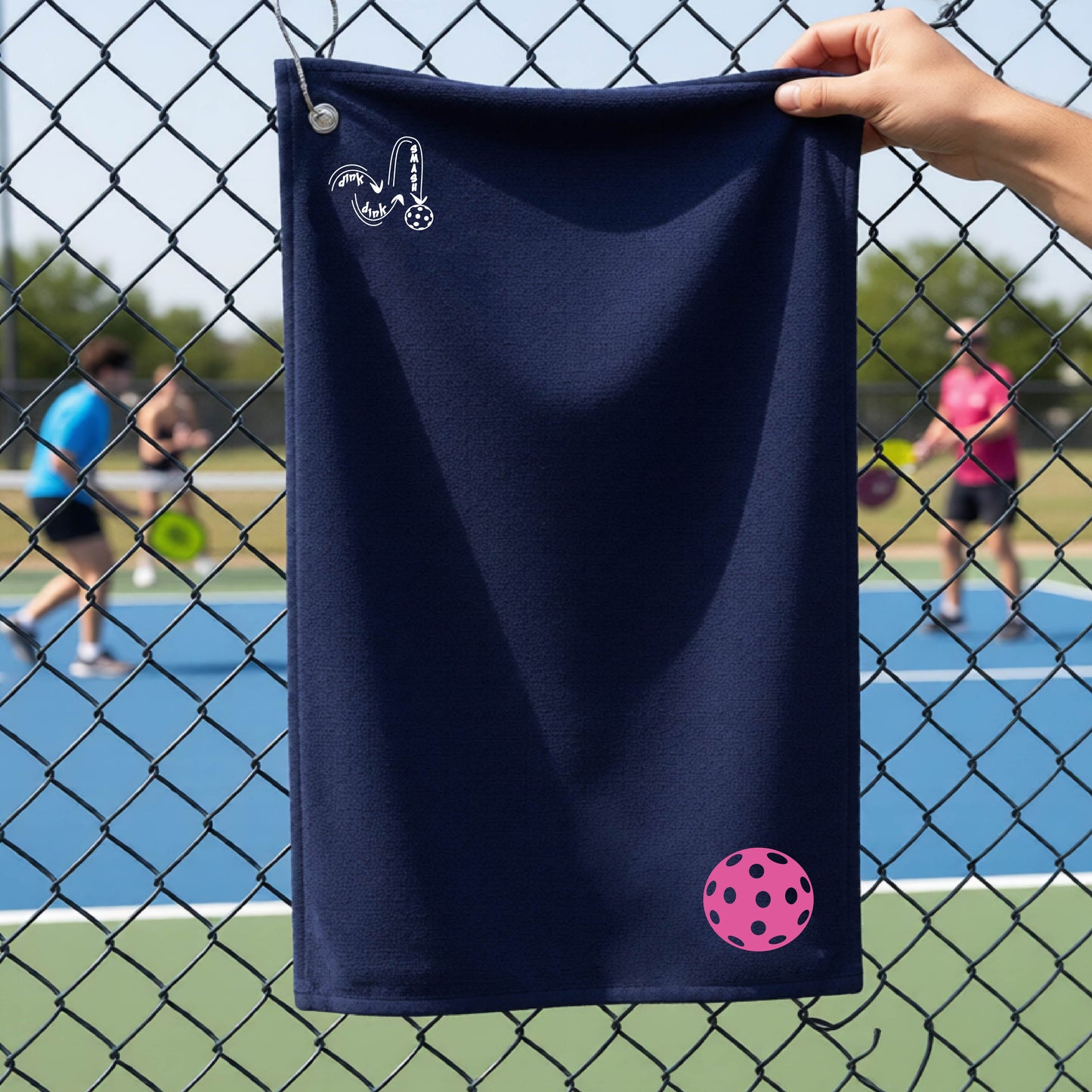A navy blue terry cloth court towel is held up in front of a chain-link fence at a pickleall court. The towel features a silver metal grommet and hook at the top, with the text"DINK DINK SMASH" and a colorful pickleball graphic near the bottom right.
