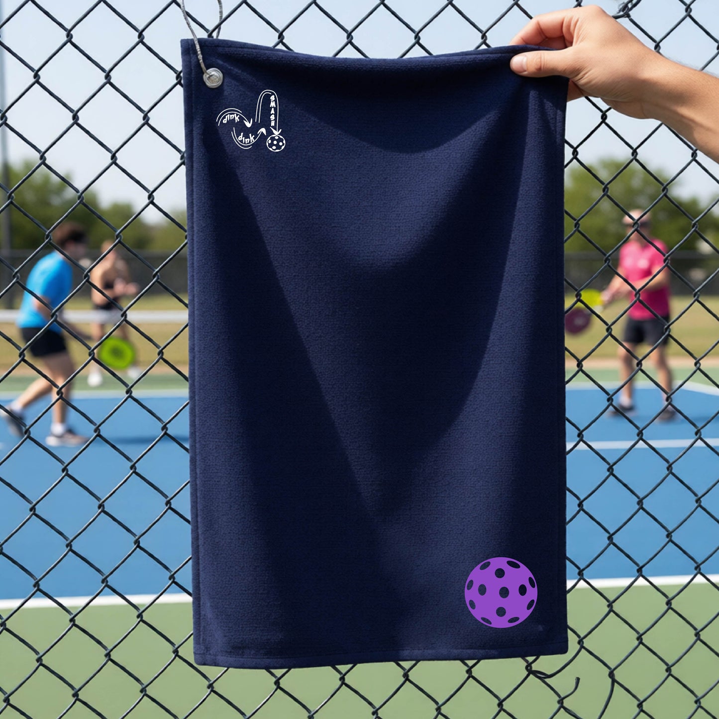 A navy blue terry cloth court towel is held up in front of a chain-link fence at a pickleall court. The towel features a silver metal grommet and hook at the top, with the text"DINK DINK SMASH" and a colorful pickleball graphic near the bottom right.