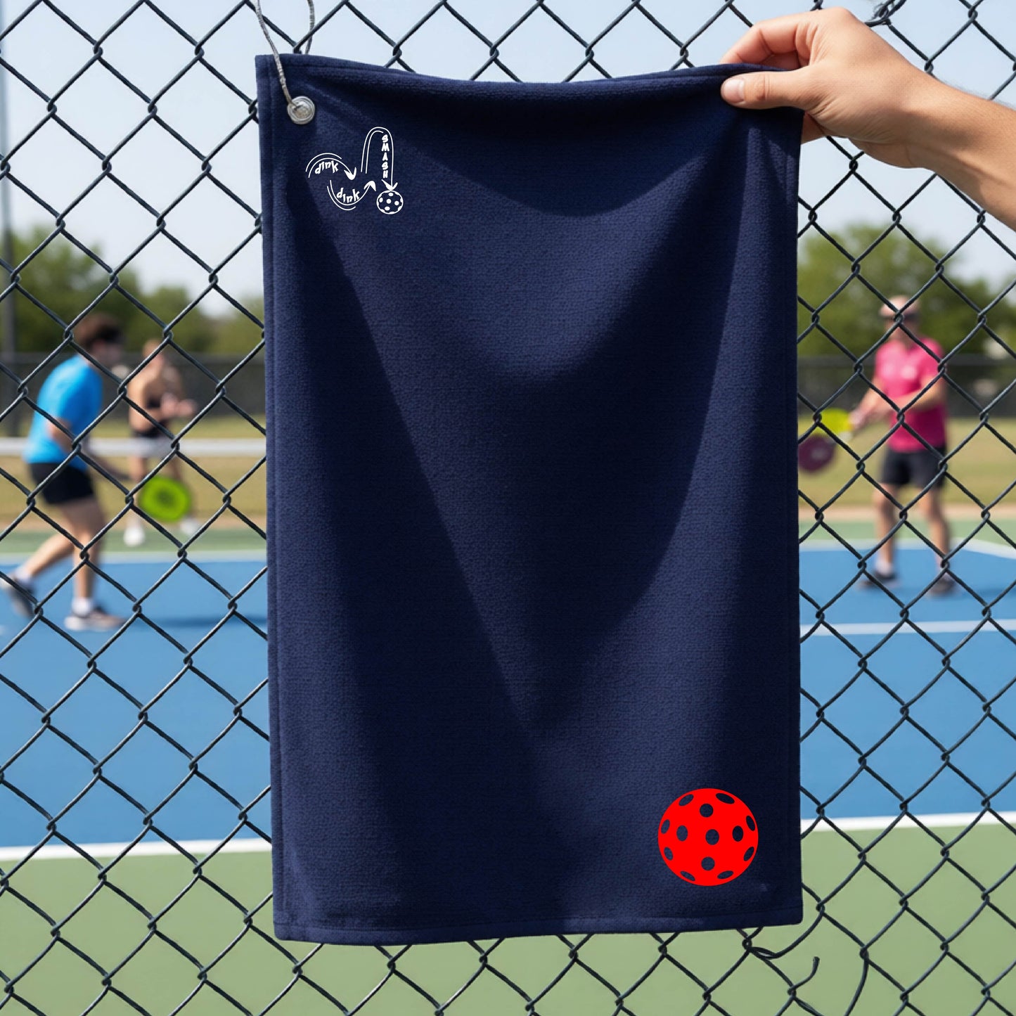 A navy blue terry cloth court towel is held up in front of a chain-link fence at a pickleall court. The towel features a silver metal grommet and hook at the top, with the text"DINK DINK SMASH" and a colorful pickleball graphic near the bottom right.