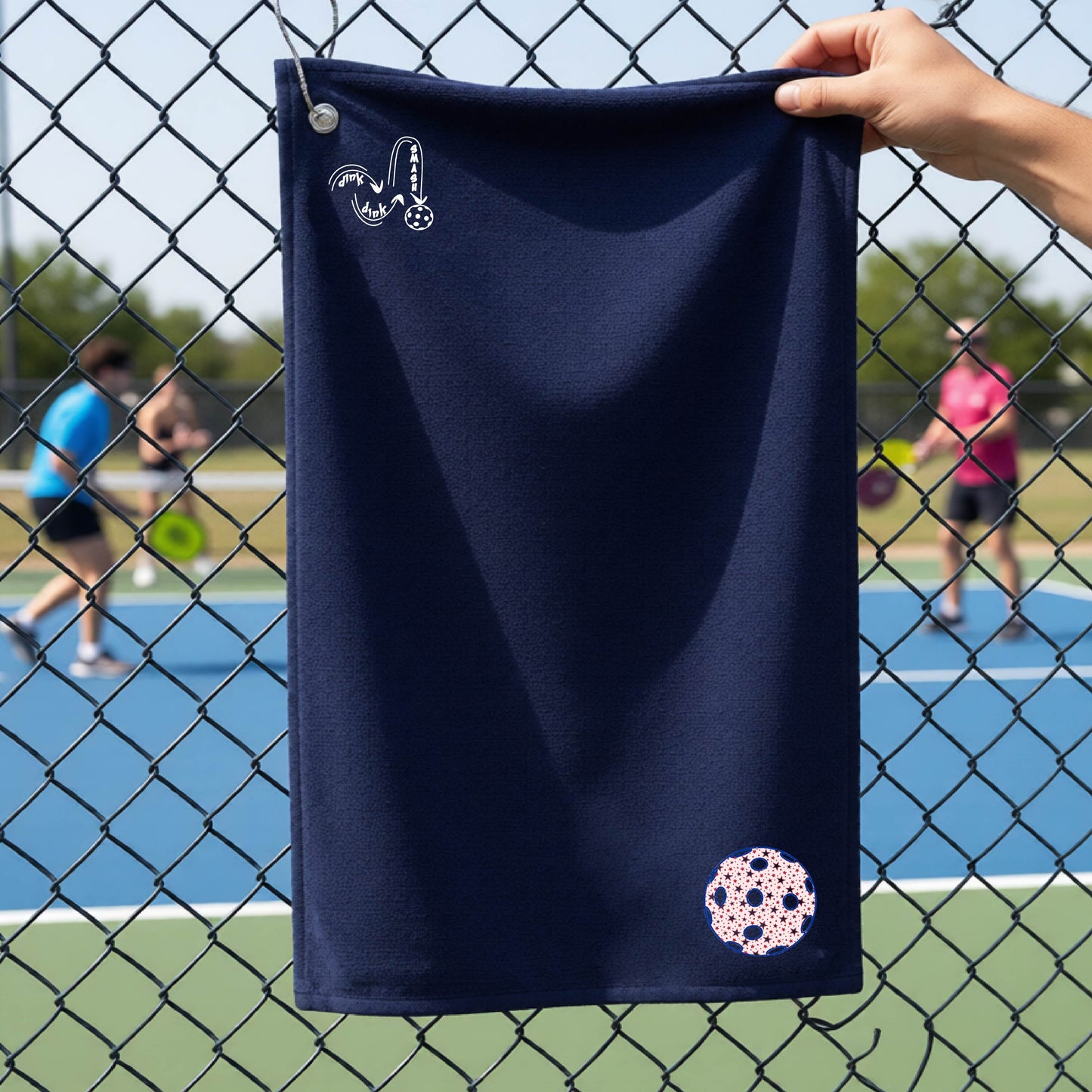 A navy blue terry cloth court towel is held up in front of a chain-link fence at a pickleall court. The towel features a silver metal grommet and hook at the top, with the text"DINK DINK SMASH" and a colorful pickleball graphic near the bottom right.