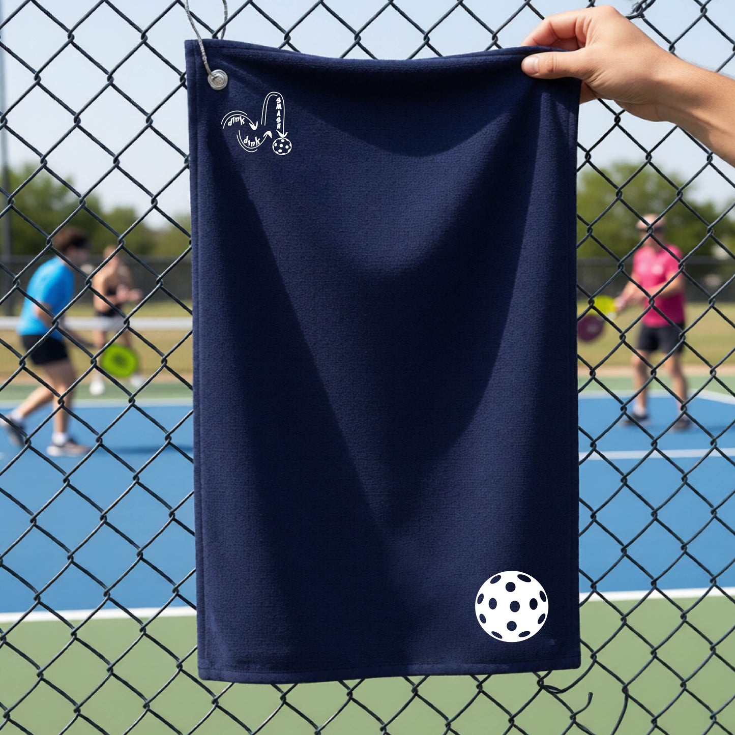 A navy blue terry cloth court towel is held up in front of a chain-link fence at a pickleall court. The towel features a silver metal grommet and hook at the top, with the text"DINK DINK SMASH" and a colorful pickleball graphic near the bottom right.