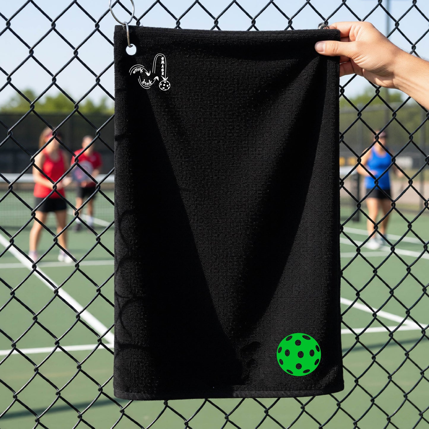 A black terry cloth towel is held up in front of a chain-link fence at a pickleall court. The towel features a silver metal grommet and hook at the top, with the text"DINK DINK SMASH" and a colorful pickleball graphic near the bottom right.