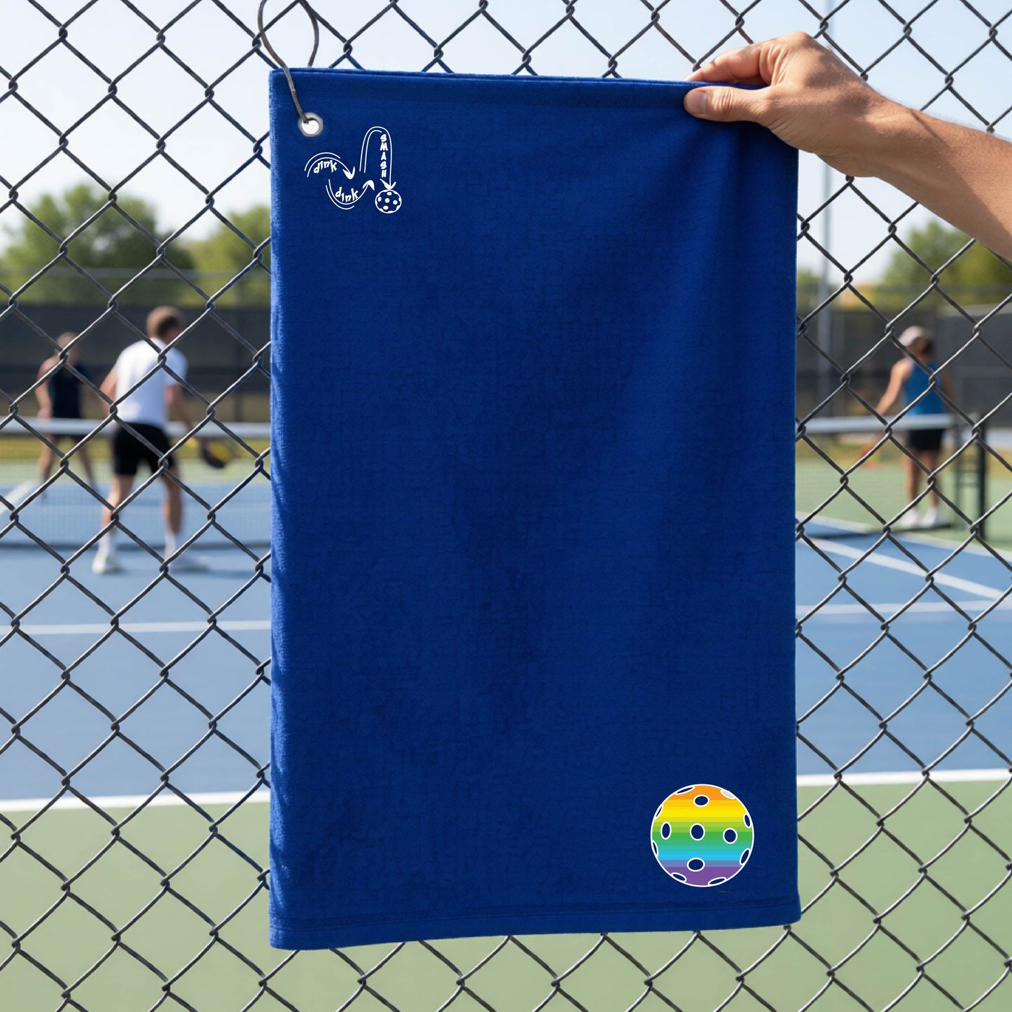 A navy blue terry cloth court towel is held up in front of a chain-link fence at a pickleall court. The towel features a silver metal grommet and hook at the top, with the text"DINK DINK SMASH" and a colorful pickleball graphic near the bottom right.