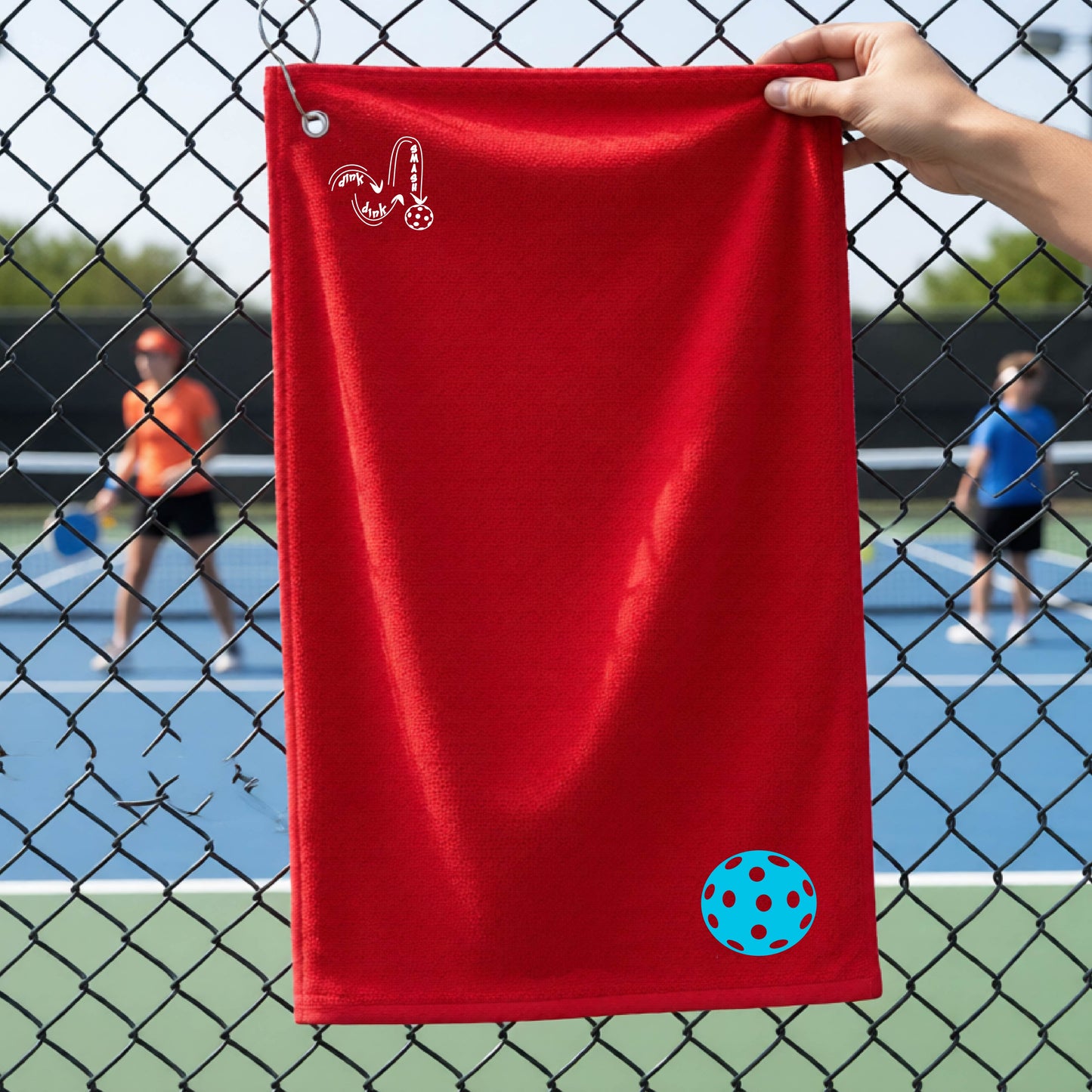 A red terry cloth court towel is held up in front of a chain-link fence at a pickleall court. The towel features a silver metal grommet and hook at the top, with the text"DINK DINK SMASH" and a colorful pickleball graphic near the bottom right.