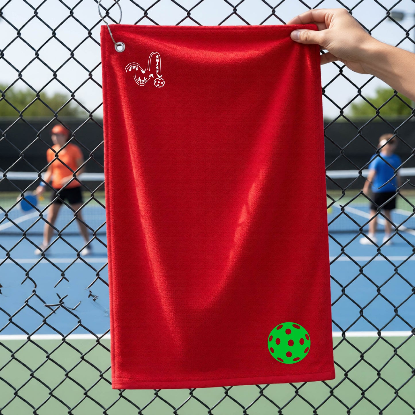 A red terry cloth court towel is held up in front of a chain-link fence at a pickleall court. The towel features a silver metal grommet and hook at the top, with the text"DINK DINK SMASH" and a colorful pickleball graphic near the bottom right.
