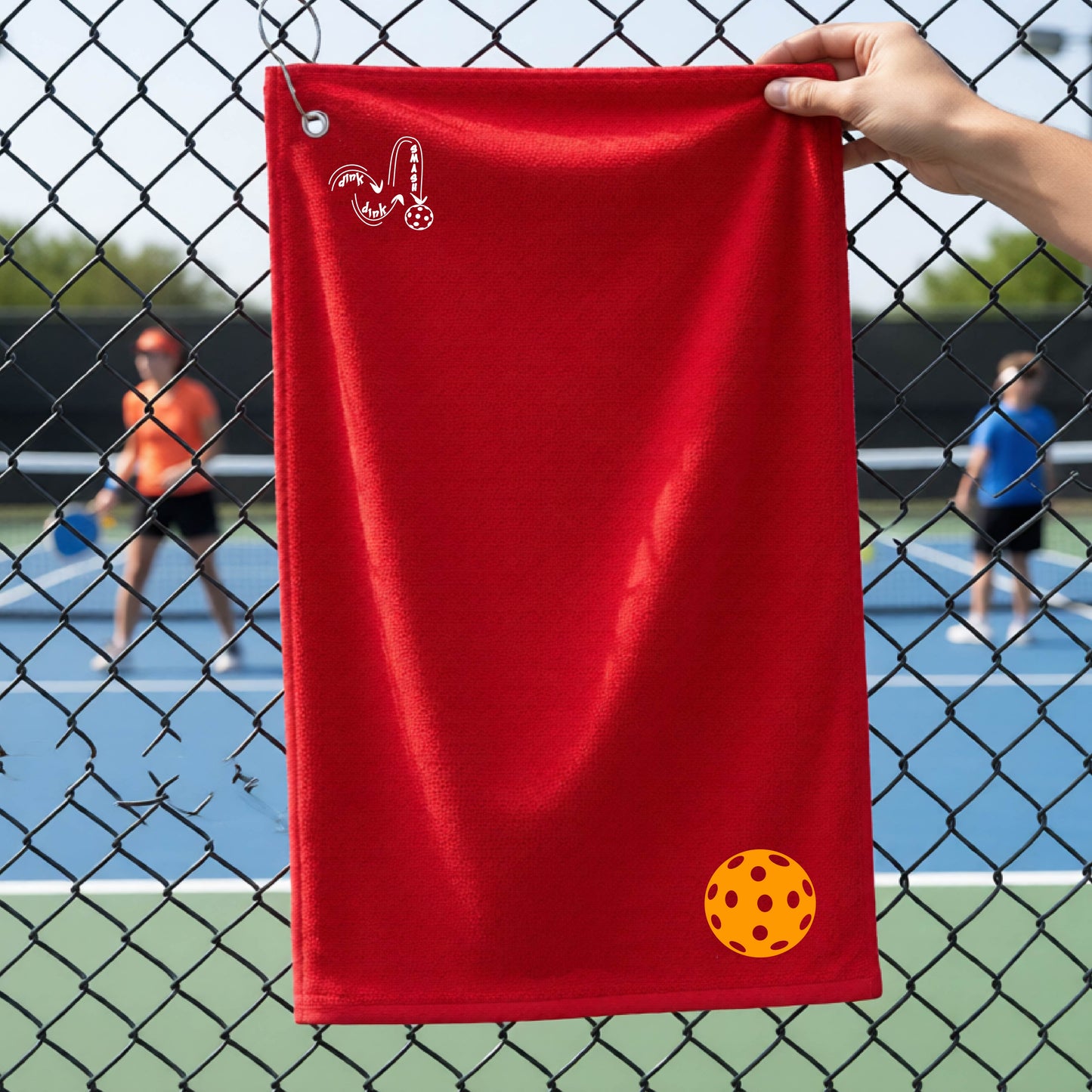 A red terry cloth court towel is held up in front of a chain-link fence at a pickleall court. The towel features a silver metal grommet and hook at the top, with the text"DINK DINK SMASH" and a colorful pickleball graphic near the bottom right.