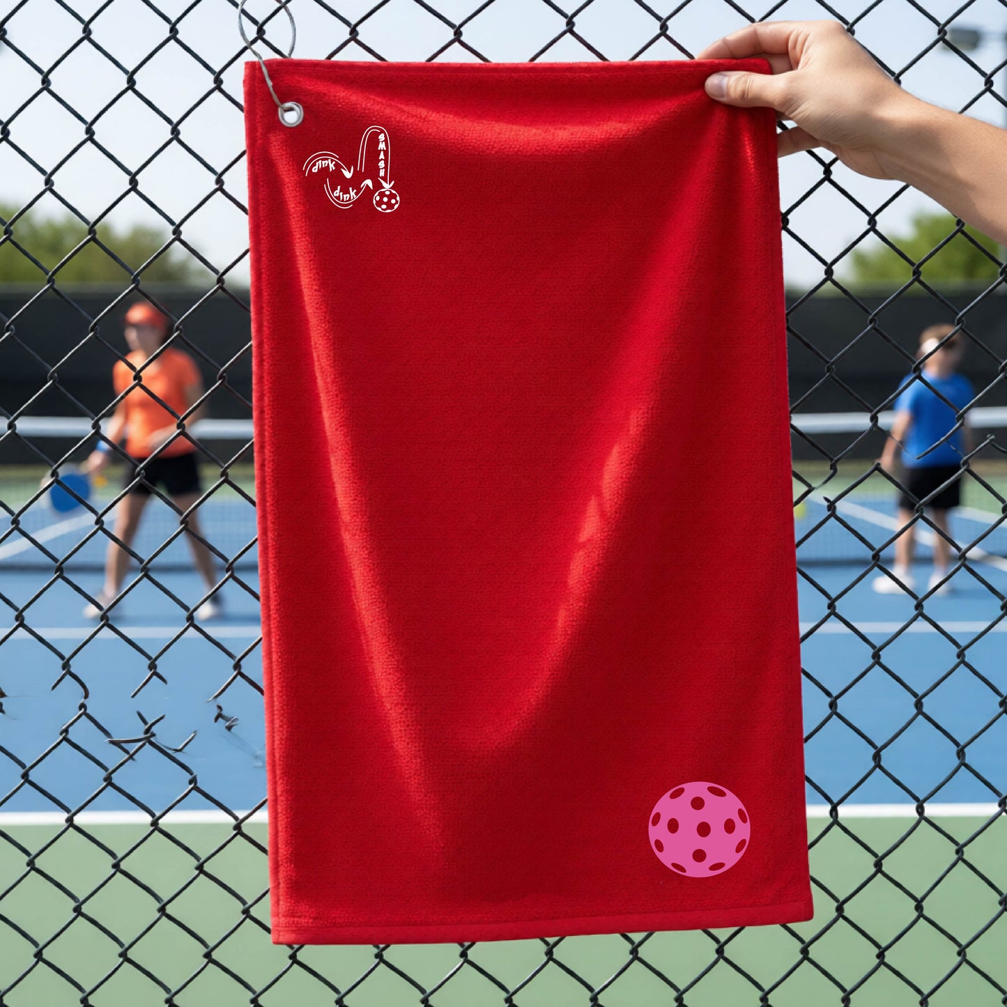 A red terry cloth court towel is held up in front of a chain-link fence at a pickleall court. The towel features a silver metal grommet and hook at the top, with the text"DINK DINK SMASH" and a colorful pickleball graphic near the bottom right.