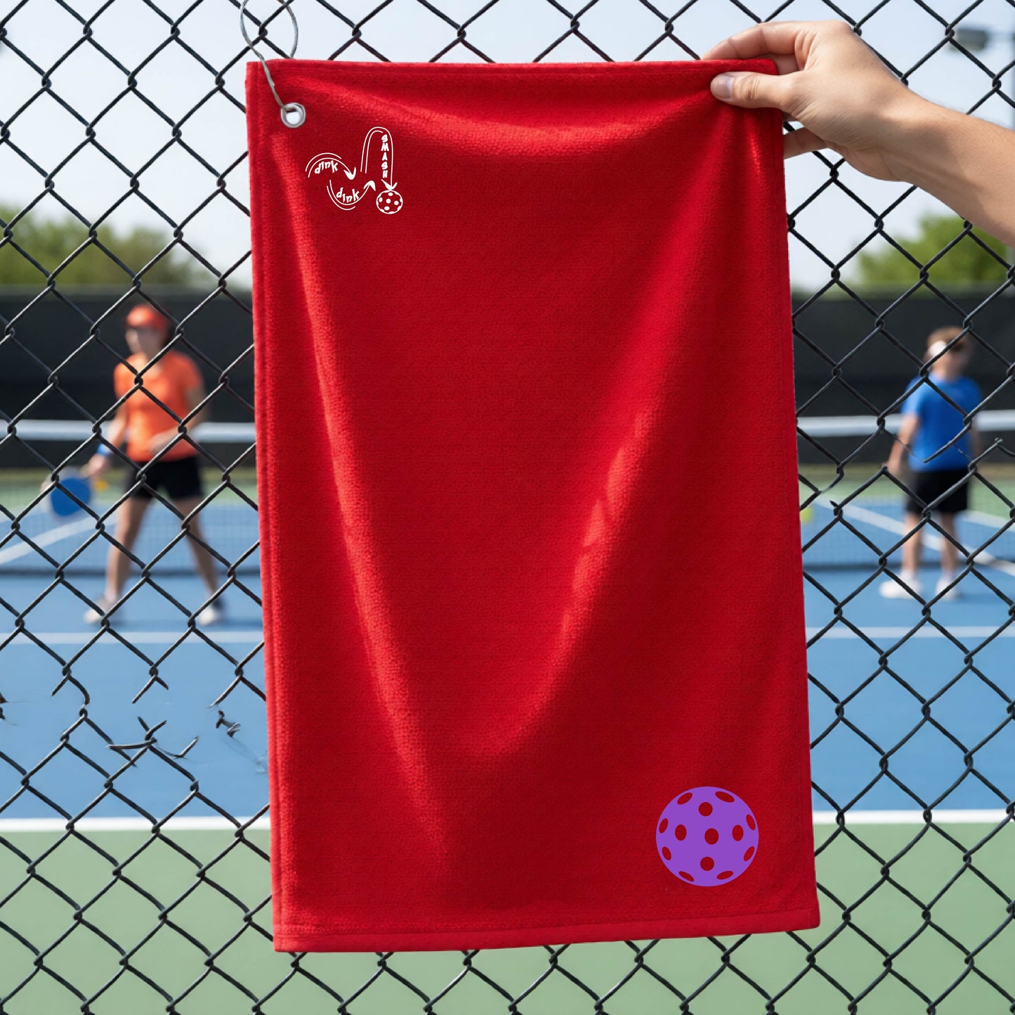 A red terry cloth court towel is held up in front of a chain-link fence at a pickleall court. The towel features a silver metal grommet and hook at the top, with the text"DINK DINK SMASH" and a colorful pickleball graphic near the bottom right.