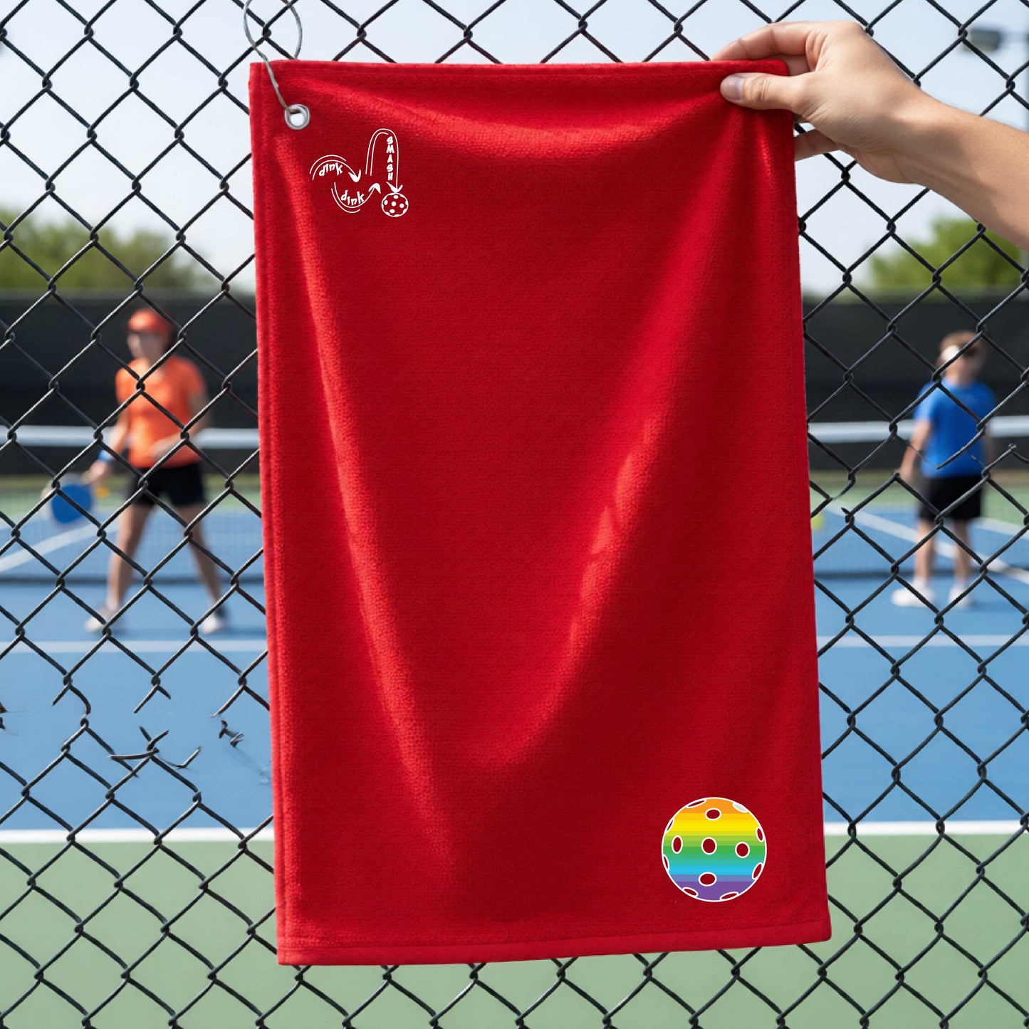 A red terry cloth court towel is held up in front of a chain-link fence at a pickleall court. The towel features a silver metal grommet and hook at the top, with the text"DINK DINK SMASH" and a colorful pickleball graphic near the bottom right.