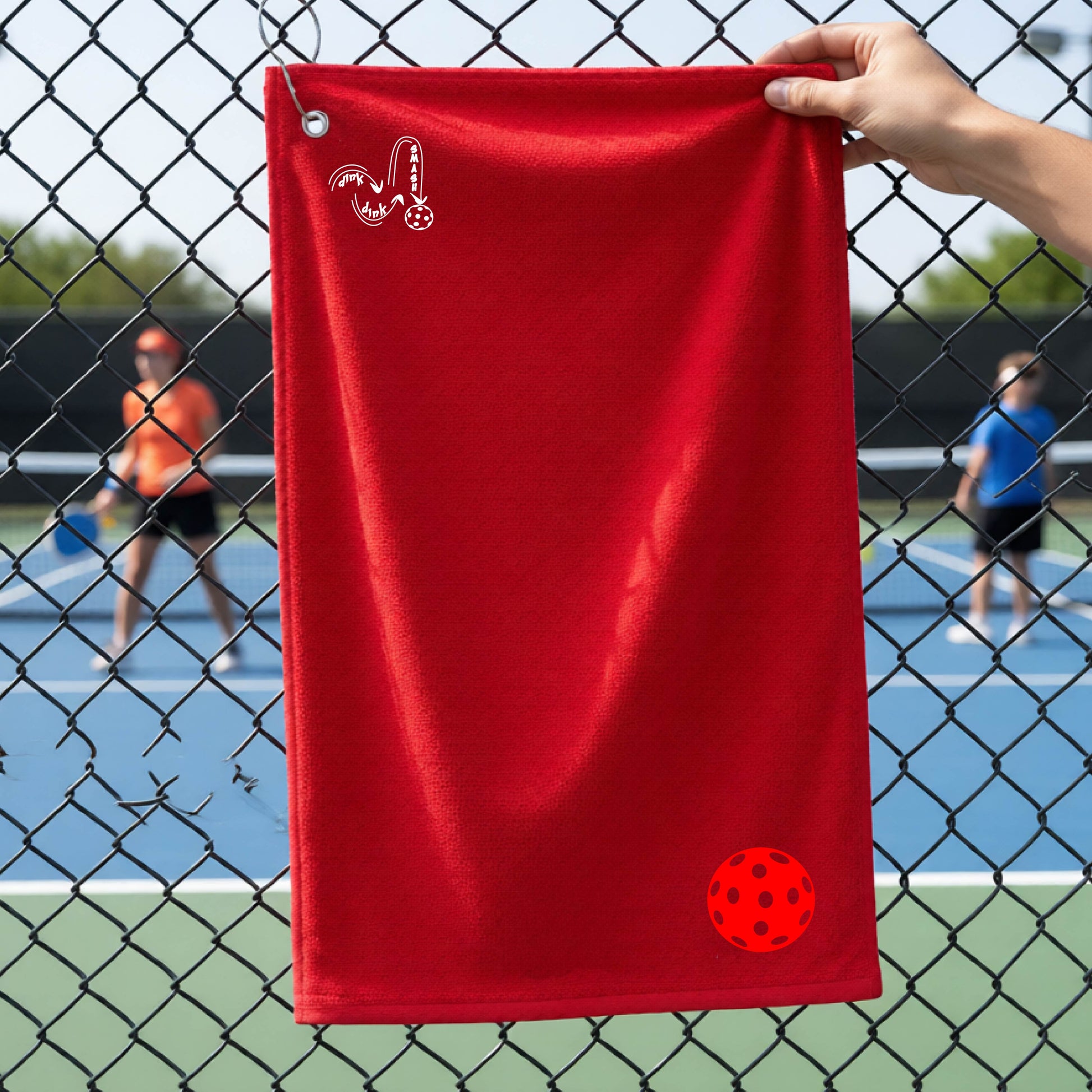 A red terry cloth court towel is held up in front of a chain-link fence at a pickleall court. The towel features a silver metal grommet and hook at the top, with the text"DINK DINK SMASH" and a colorful pickleball graphic near the bottom right.