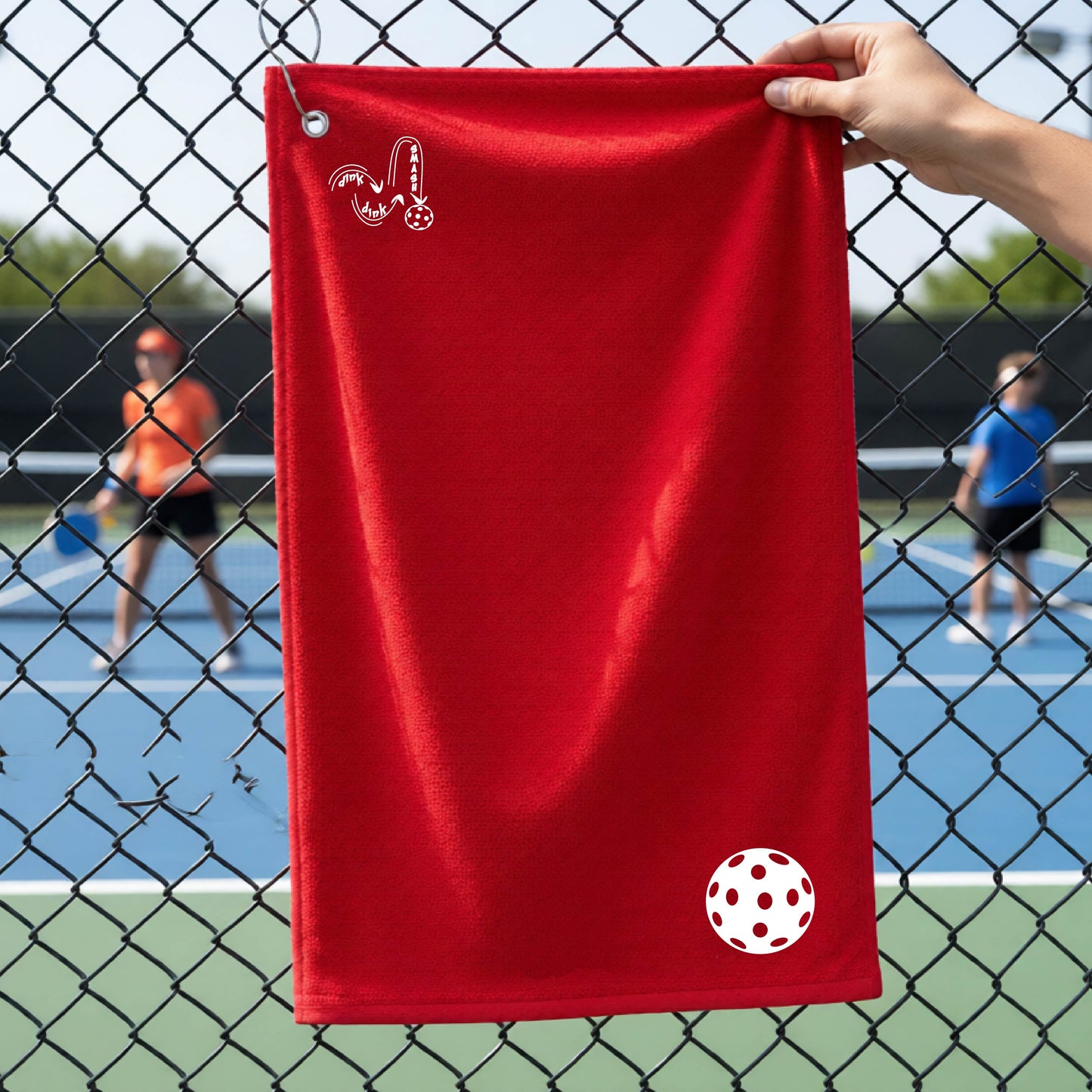 A red terry cloth court towel is held up in front of a chain-link fence at a pickleall court. The towel features a silver metal grommet and hook at the top, with the text"DINK DINK SMASH" and a colorful pickleball graphic near the bottom right.