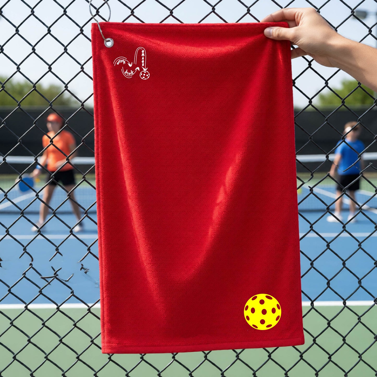 A red terry cloth court towel is held up in front of a chain-link fence at a pickleall court. The towel features a silver metal grommet and hook at the top, with the text"DINK DINK SMASH" and a colorful pickleball graphic near the bottom right.