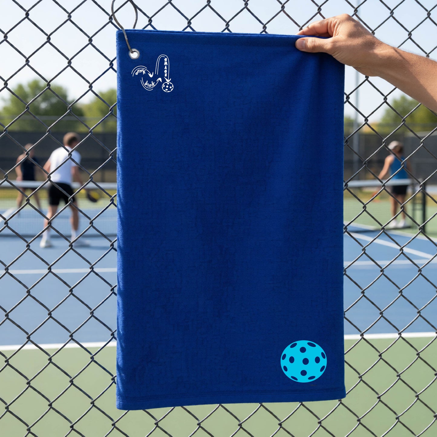 A royal blue terry cloth court towel is held up in front of a chain-link fence at a pickleall court. The towel features a silver metal grommet and hook at the top, with the text"DINK DINK SMASH" and a colorful pickleball graphic near the bottom right.