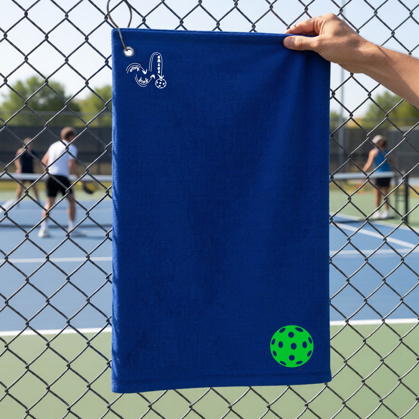 A royal blue terry cloth court towel is held up in front of a chain-link fence at a pickleall court. The towel features a silver metal grommet and hook at the top, with the text"DINK DINK SMASH" and a colorful pickleball graphic near the bottom right.