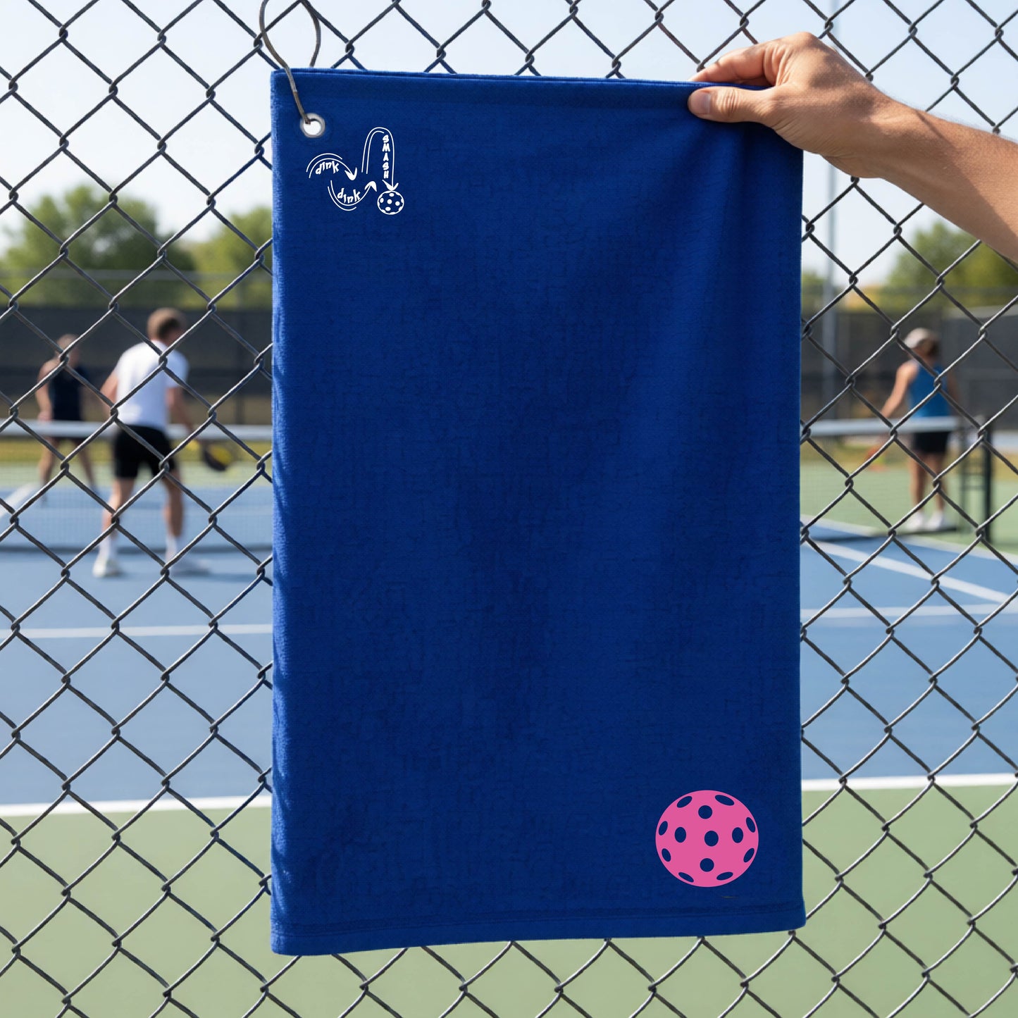 A royal blue terry cloth court towel is held up in front of a chain-link fence at a pickleall court. The towel features a silver metal grommet and hook at the top, with the text"DINK DINK SMASH" and a colorful pickleball graphic near the bottom right.