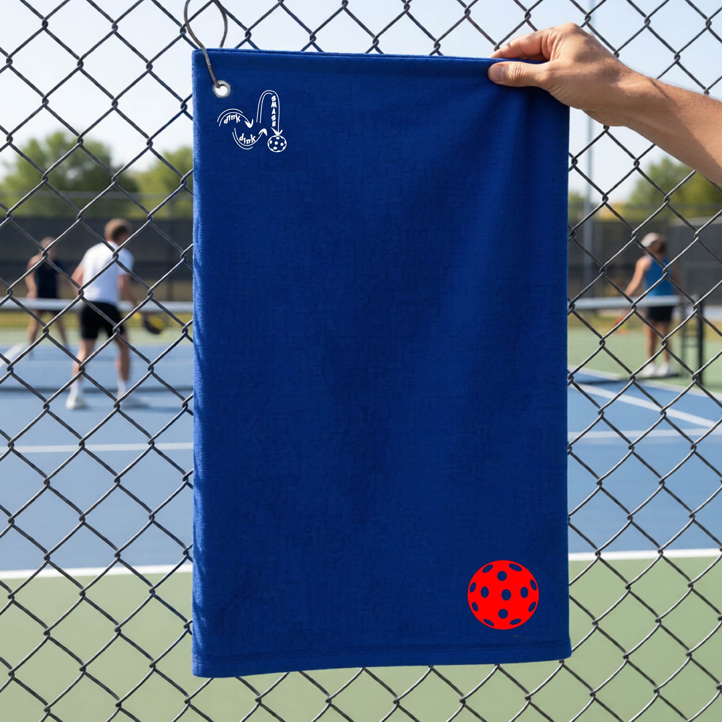 A royal blue terry cloth court towel is held up in front of a chain-link fence at a pickleall court. The towel features a silver metal grommet and hook at the top, with the text"DINK DINK SMASH" and a colorful pickleball graphic near the bottom right.